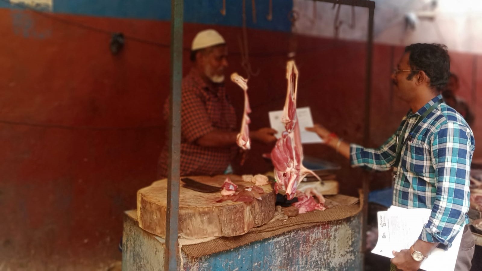 man in checked short speaking to a man handling raw meat